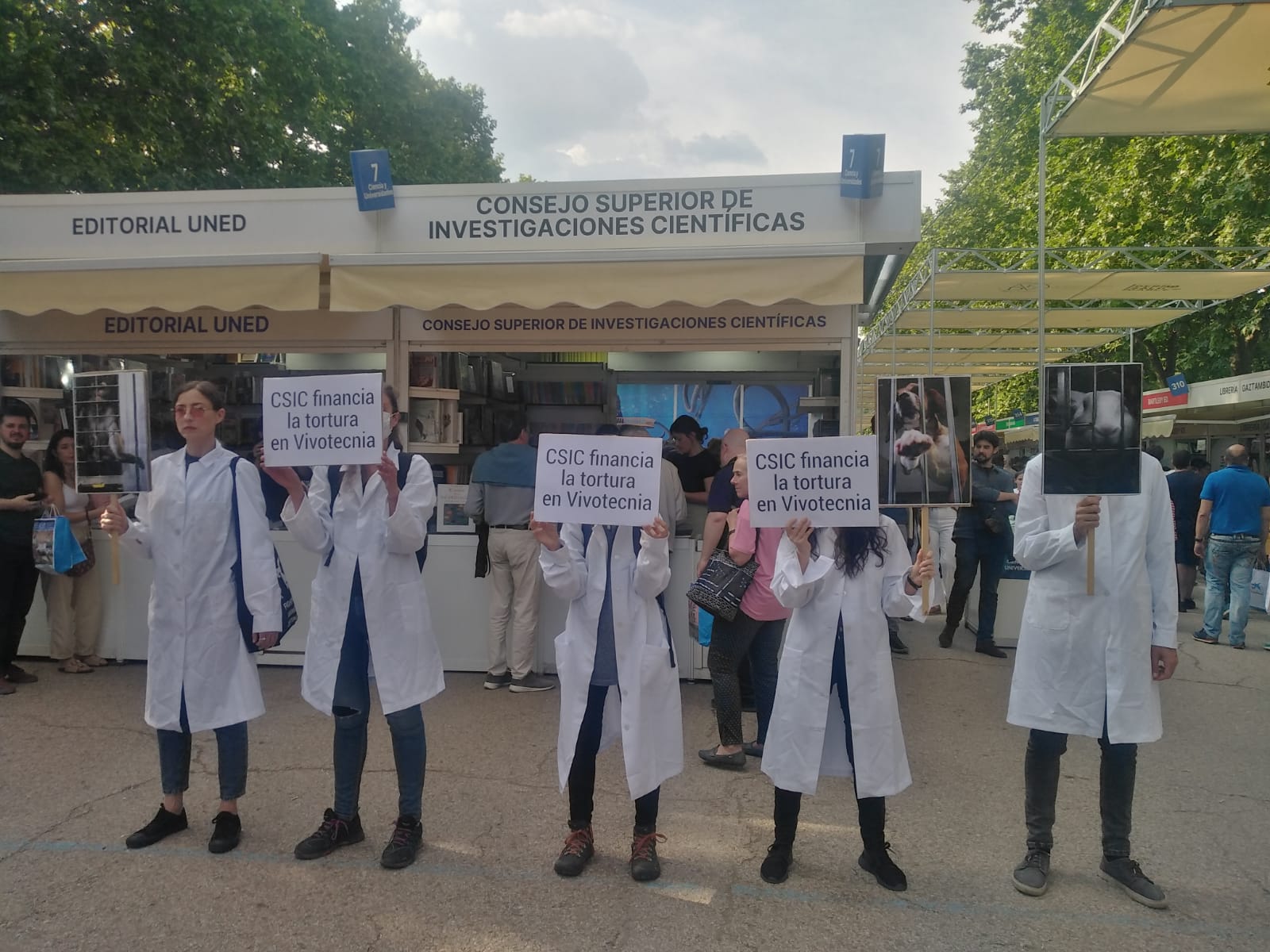 Fila de activistas con batas blancas y carteles denunciando la colaboración entre el CSIV y Vivotecnia, delante del puesto del CSIC en la Feria del Libro de Madrid.